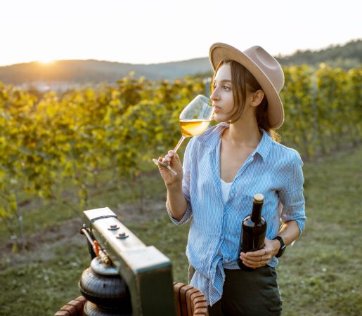 Young and cheerful woman tasting wine, while standing near the press machine on the vineyard on a sunny evening